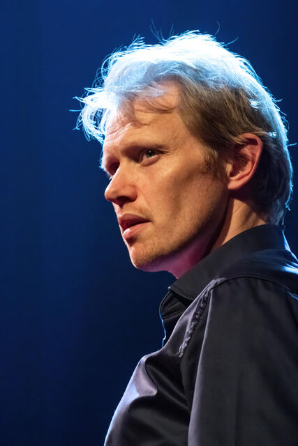 Portret van een man met lichtblond haar, die serieus kijkt naar de rechterkant, tegen een blauwe achtergrond.