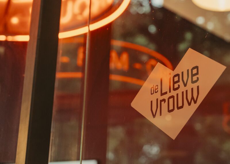 Afbeelding van een etiket met de tekst "de Lieve Vrouw" op een glazen deur, met een warme achtergrondverlichting.