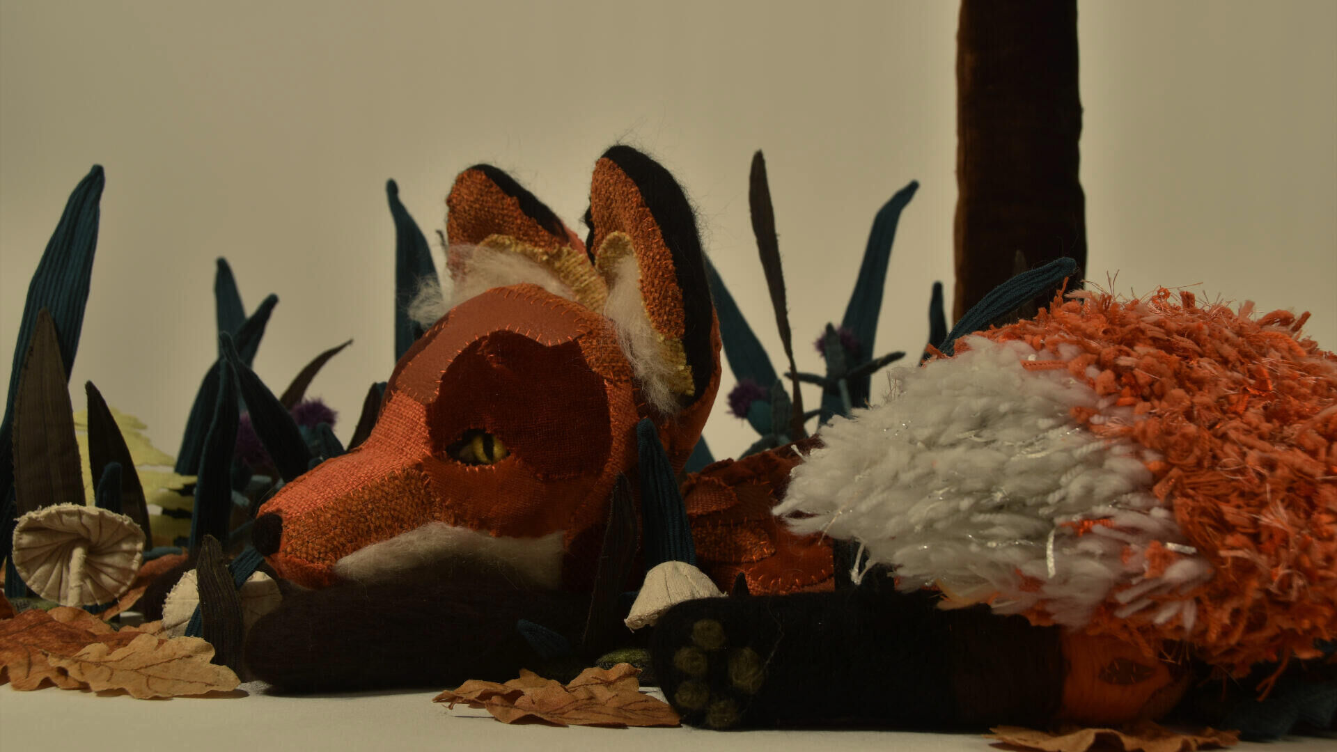 Een handgemaakte vos van stof, omringd door kleurrijke bladeren en gras in een natuurlijke setting.