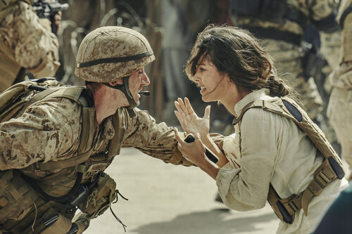 Soldaat en vrouw ontmoeten elkaar met blijdschap in een oorlogsgebied, omgeven door andere soldaten.