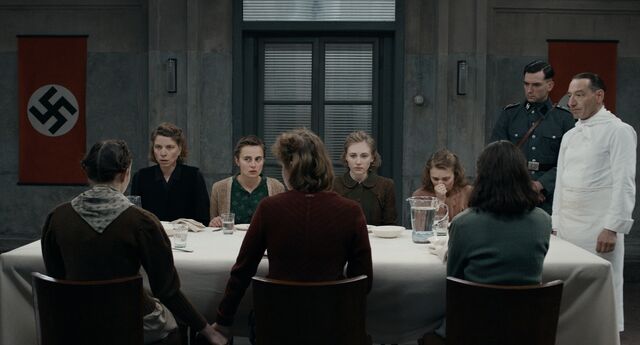 Een groep mensen zit aan een tafel in een donkere ruimte, terwijl figuren in uniform toekijken.