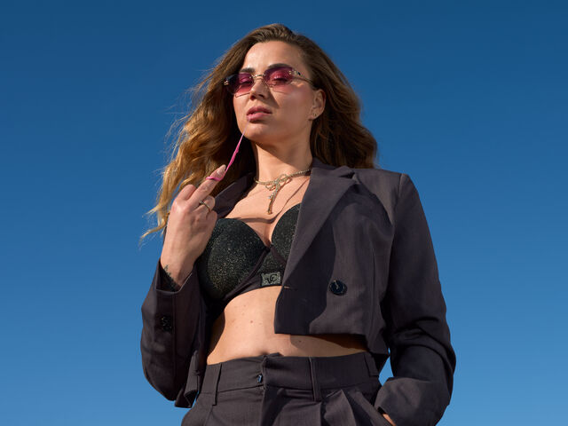 Model in een zwarte outfit met een blazer en zonnebril, poseert tegen een blauwe lucht.
