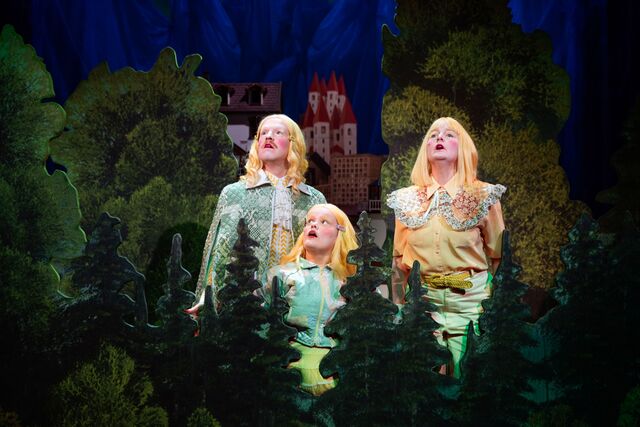 Drie actrices met lange blonde haar, gekleed in groene kostuums, zingen midden in een bosachtige setting.