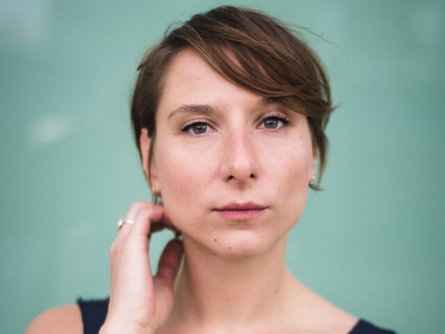 Portret van een vrouw met kort bruin haar en een neutrale gezichtsuitdrukking, tegen een groene achtergrond.
