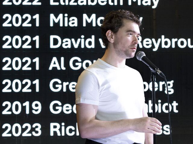 Man in witte T-shirt staat op een podium met microfoon, terwijl namen en jaartallen op een scherm achter hem worden weergegeven.