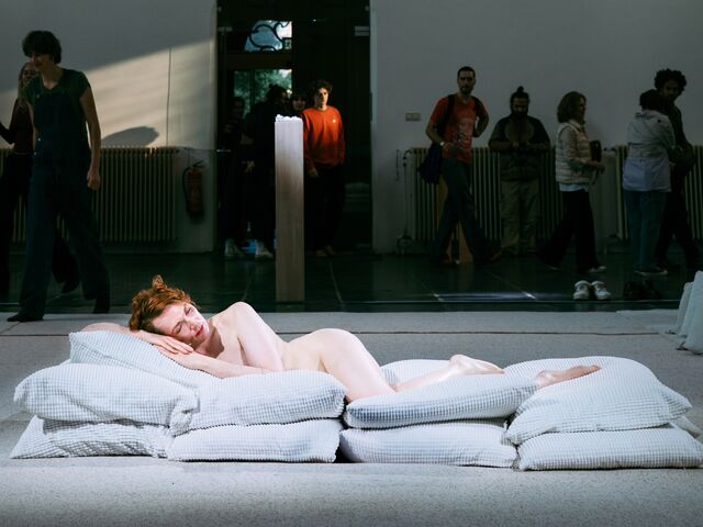 Een naakte vrouw ligt op een stapel kussens in een museum, terwijl bezoekers op de achtergrond rondlopen.