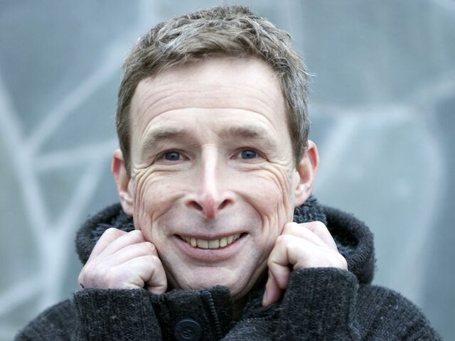 Een man met een korte, grijze haarsnit glimlacht terwijl hij zijn handen onder zijn kin houdt, gekleed in een zwarte jas.