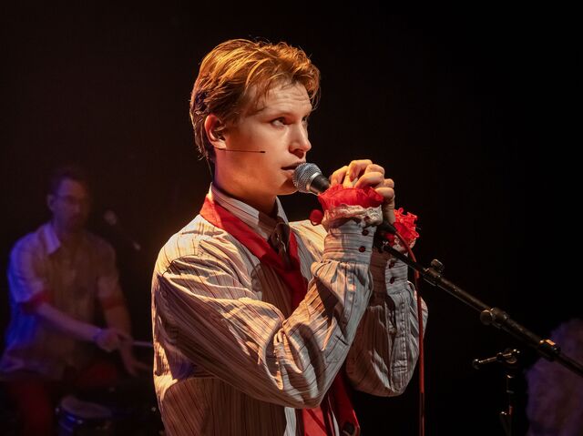 Jongeman met een microfoon op een podium, zingend, met een band op de achtergrond.