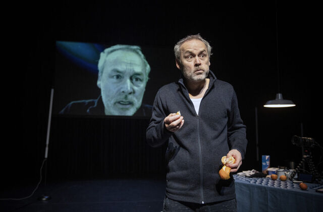 Man in zwarte kleding staat op het podium met een sinaasappel in zijn hand, terwijl een groot scherm zijn gezicht toont.