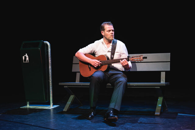 Scenefoto van Martijn Koning die gitaar speelt op een bankje met een afvalbak op de achtergrond, in een donkere theateromgeving.