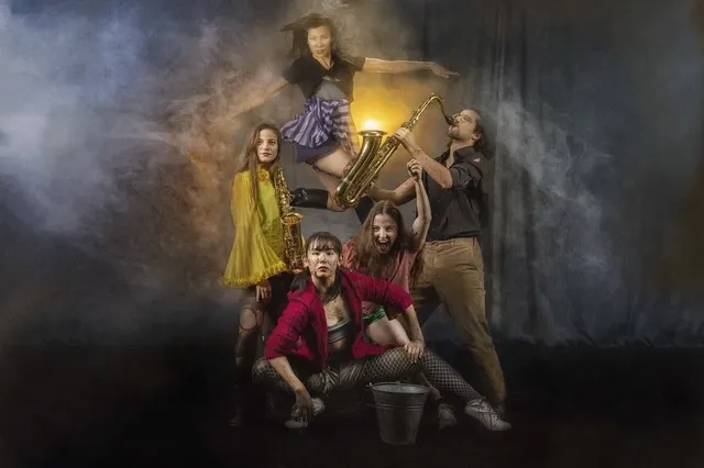 Een groep van vijf mensen poseert dramatisch op een gestapelde set van meubels in een rokerige achtergrond.