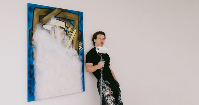 Bram Raven Kangee Ravenhorst leunt tegen een muur naast een abstracte schilderij, gekleed in een zwart shirt en met verfsporen op de broek.