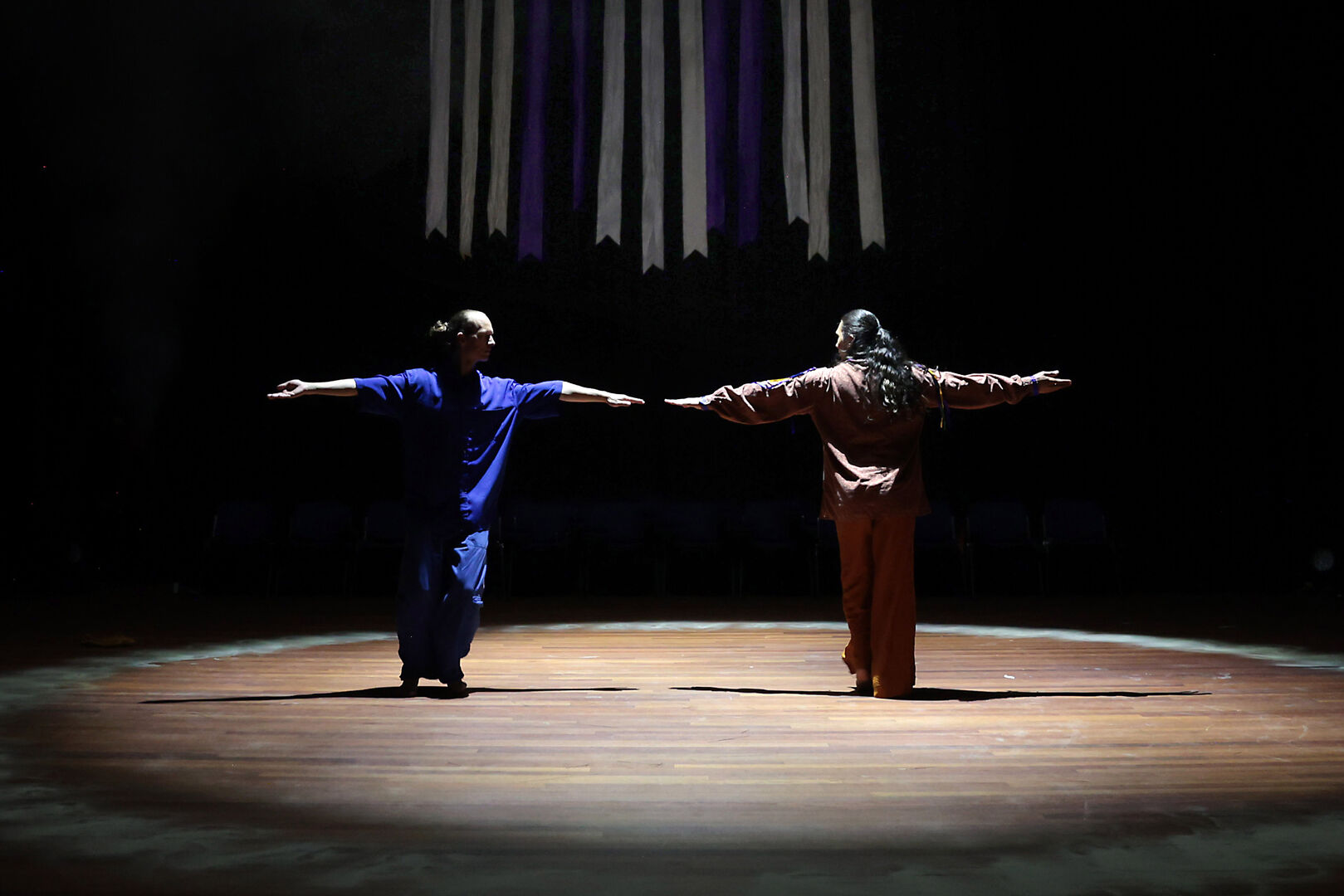 Twee dansers staan met hun rug naar de camera, met uitgestrekte armen op een verlichte houten vloer in een donkere omgeving.