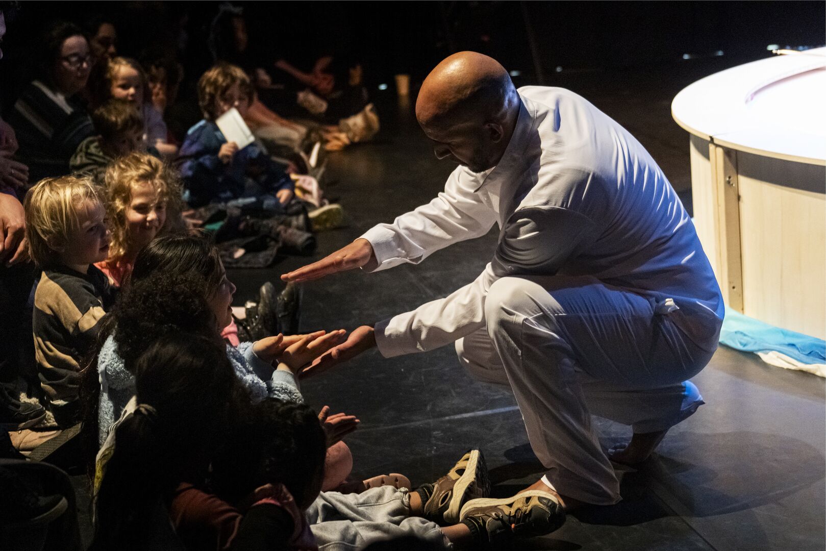 Achref Adhadhi in witte kleding leunt naar het publiek en reikt een hand naar een kind in de zaal.