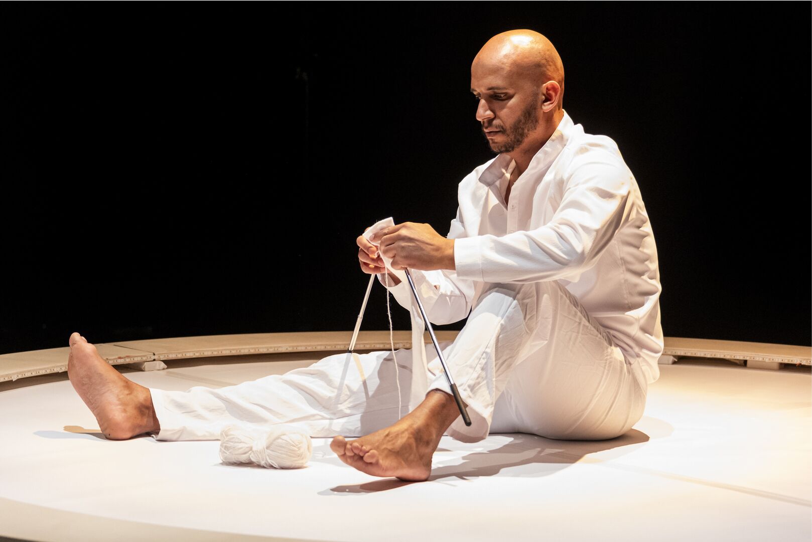 Een man in een witte outfit zit op een ronde vloer en knoopt touwen aan elkaar in een theatrale setting.