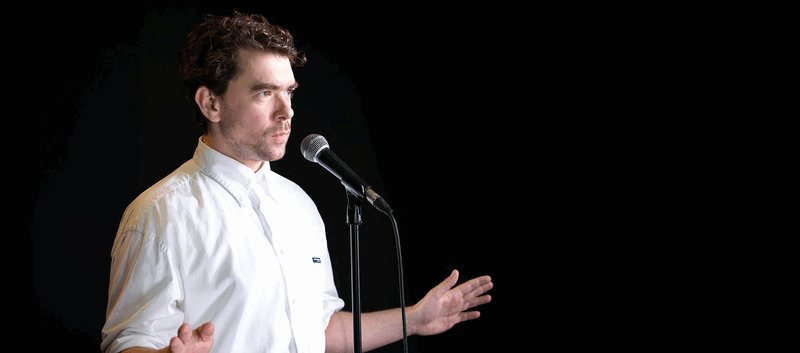 Man met een microphone die een presentatie geeft, met een zwarte achtergrond.