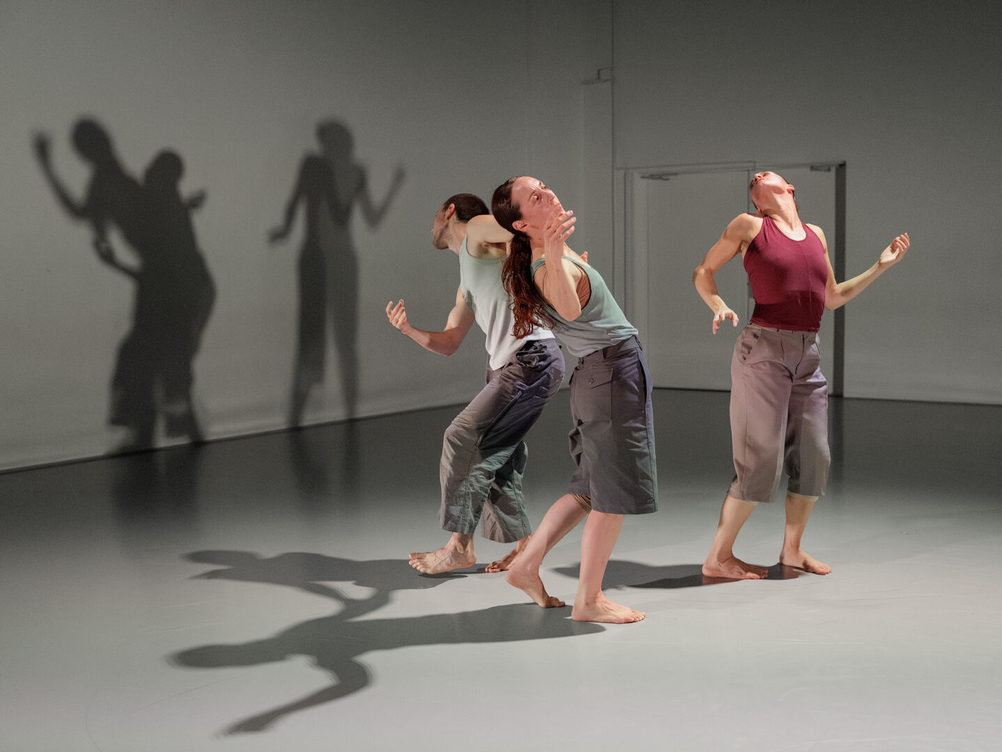 Drie dansers in losse kleding bewegen zich op een houten vloer, met schaduwen die als achtergronden zichtbaar zijn.
