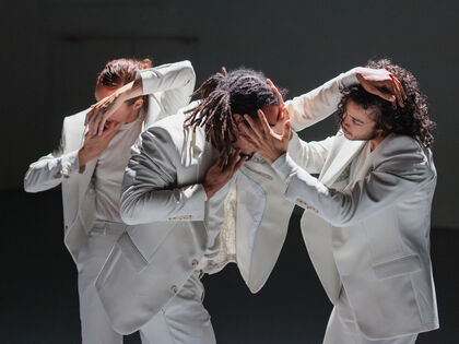 Drie dansers in witte kostuums vormen een dynamische compositie met hun handen op elkaars hoofden, tegen een neutrale achtergrond.