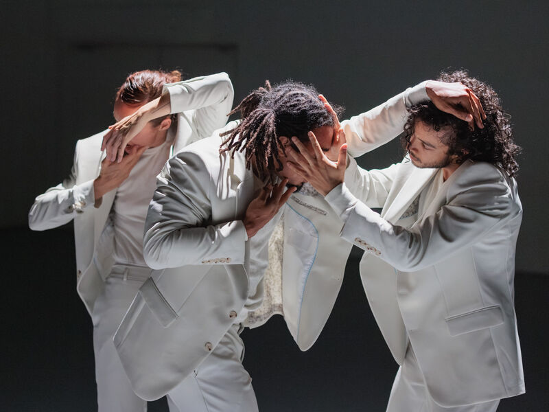 Drie dansers in witte kostuums vormen een dynamische compositie met hun handen op elkaars hoofden, tegen een neutrale achtergrond.