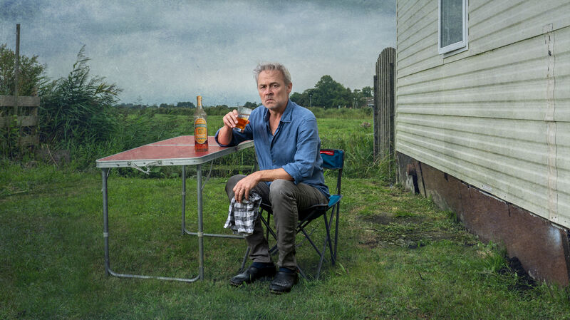 Man zit op een stoel aan een tafel buiten, met een drankje, tegen een achtergrond van een woning en onbewolkte lucht.