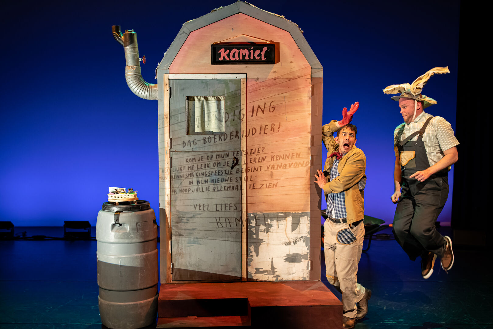 Twee acteurs voeren een sketch op bij een podium met een groot, decoratief huisje en een varkenskop op een van hen.