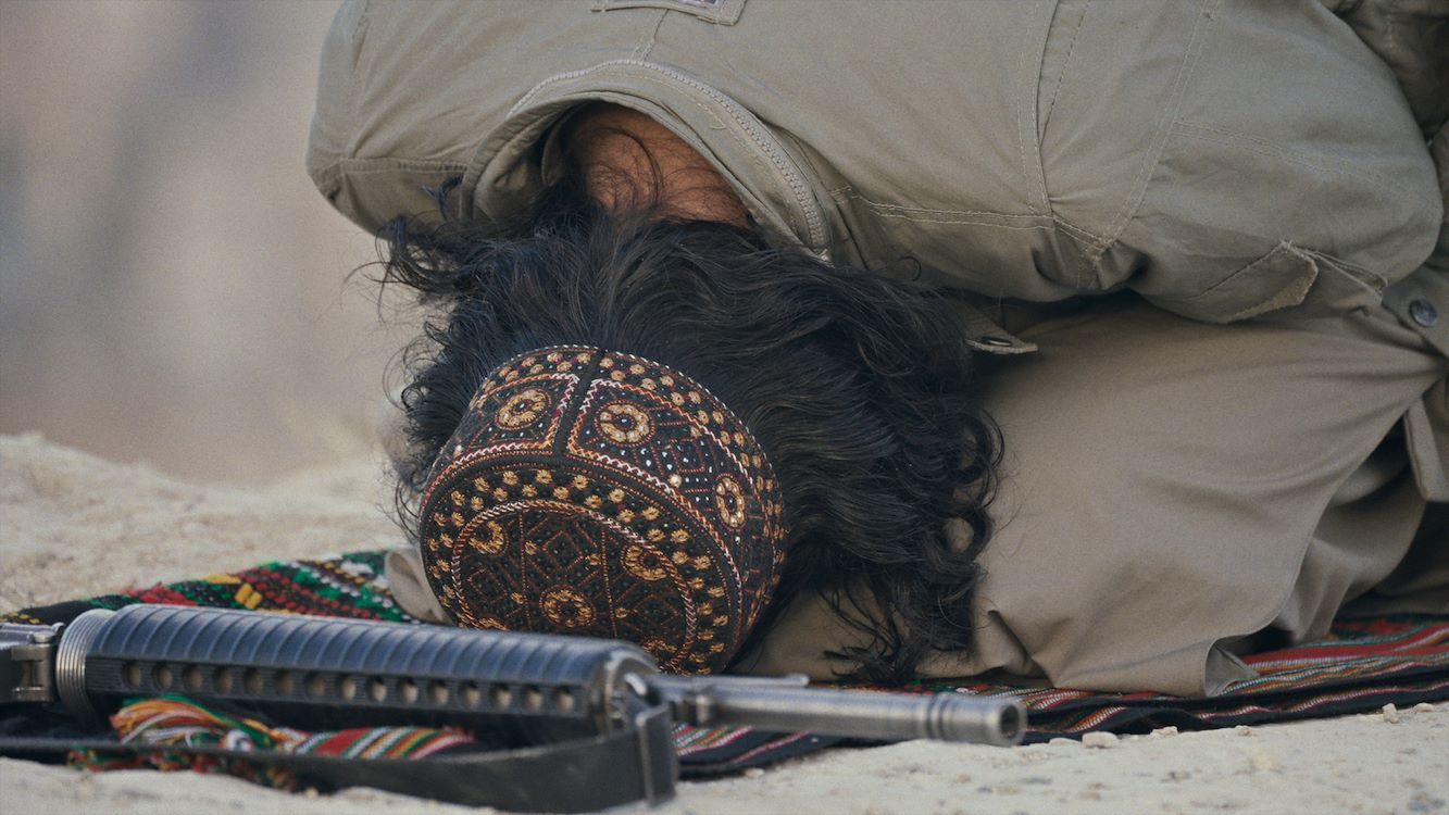 Een persoon met een hoofddoek ligt op de grond, met een geweer naast zich.