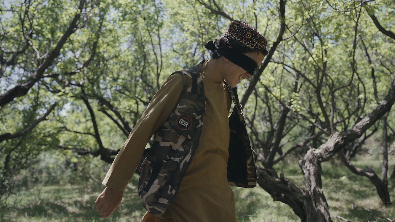 Jongeman met een blinddoek, gekleed in camouflage, loopt door een boomgaard.