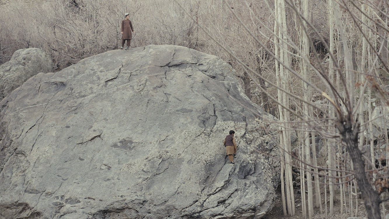 Twee mensen klimmen op een grote rots in een bossige omgeving met kale bomen.