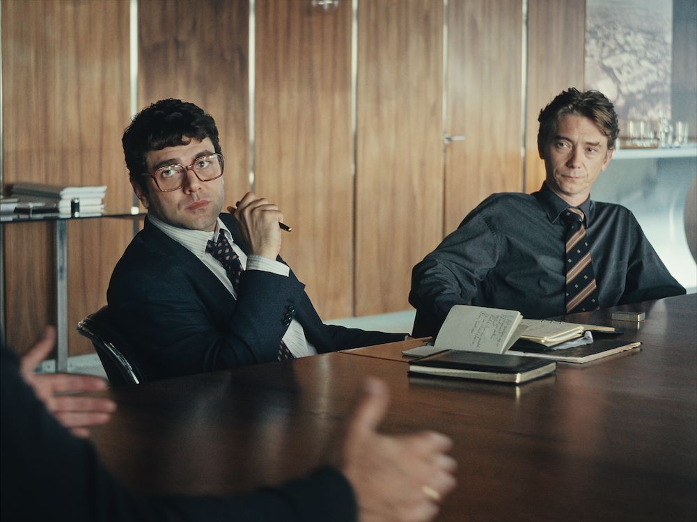 Twee mannen in een zakelijke setting, zittend aan een vergadertafel, in gesprek. Één heeft een pen in de hand.