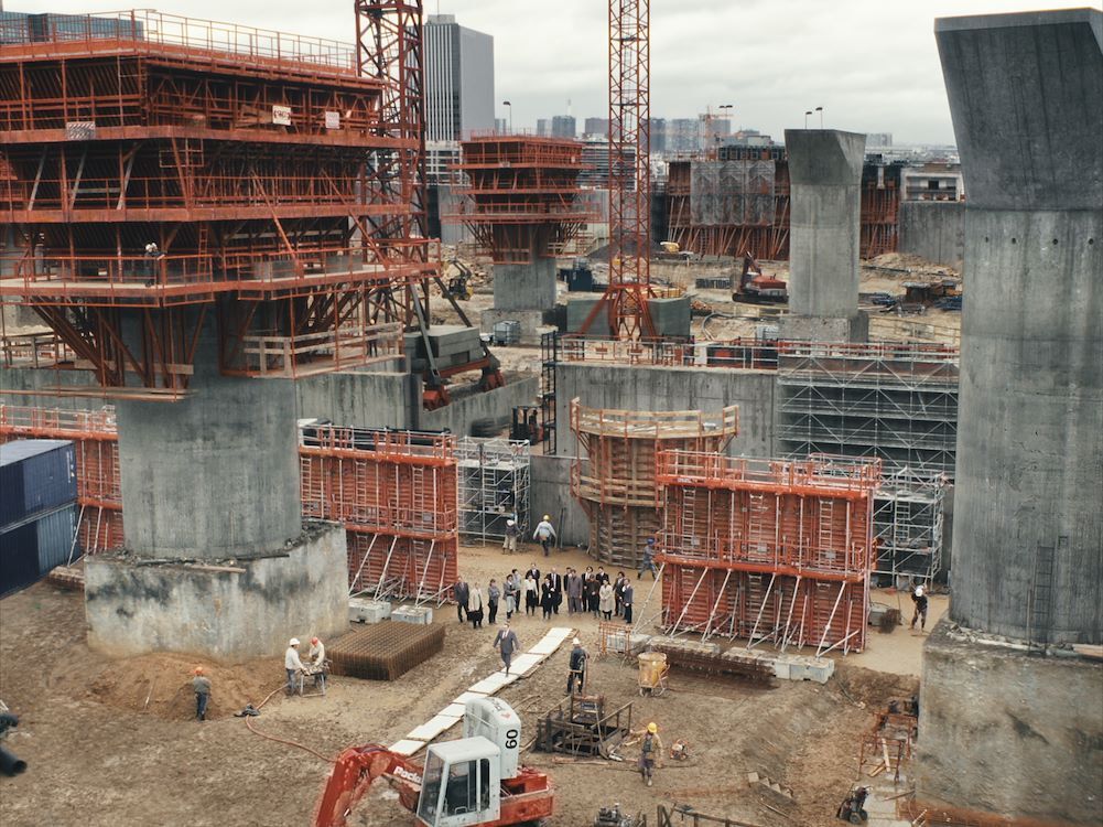 Bouwplaats met betonpilaren en steigers, verschillende machines en arbeiders aan het werk.