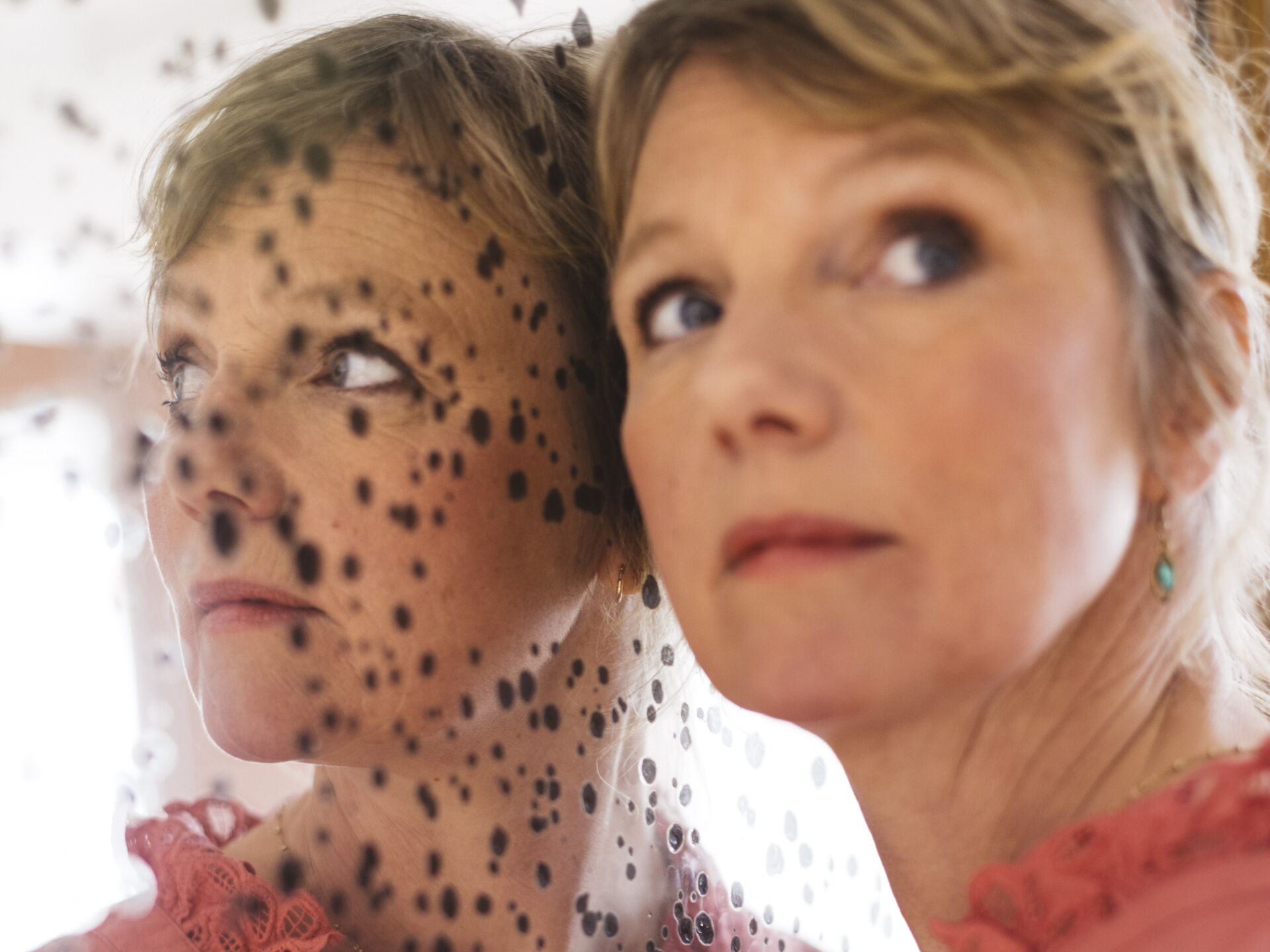 Laura van Dolron in een roze blouse kijkt contemplatief naar zichzelf in een spiegel met vlekken, met een focus op haar gelaatsuitdrukking.