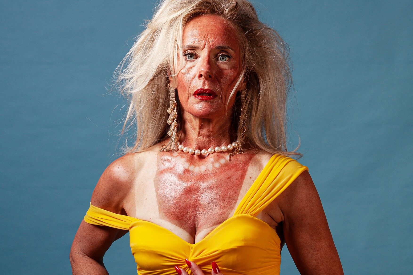 Een vrouw met lang, grijs haar, in een gele jurk en opvallende sieraden, poseert tegen een blauwe achtergrond.