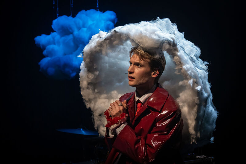 Toneelstuk met man in rode jas en wolken achtergrond, die nadenkend het publiek aankijkt.