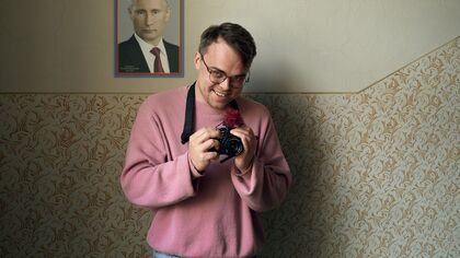 Man in een roze trui met camera, poseert voor een muur met een afbeelding van een formele man.