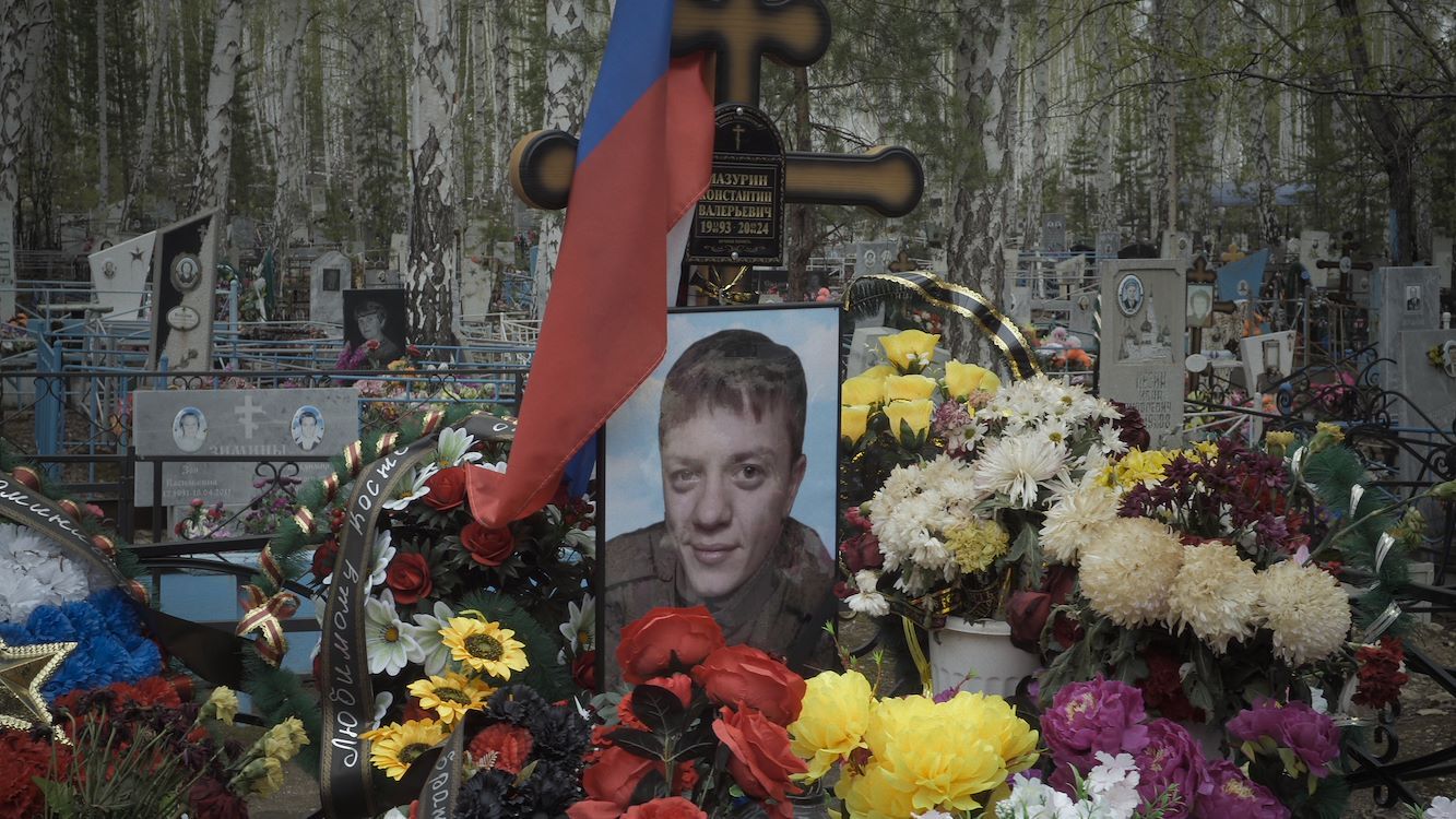 Een graf met bloemen en een foto van een persoon, omringd door bomen en een kruis, met een Russische vlag.