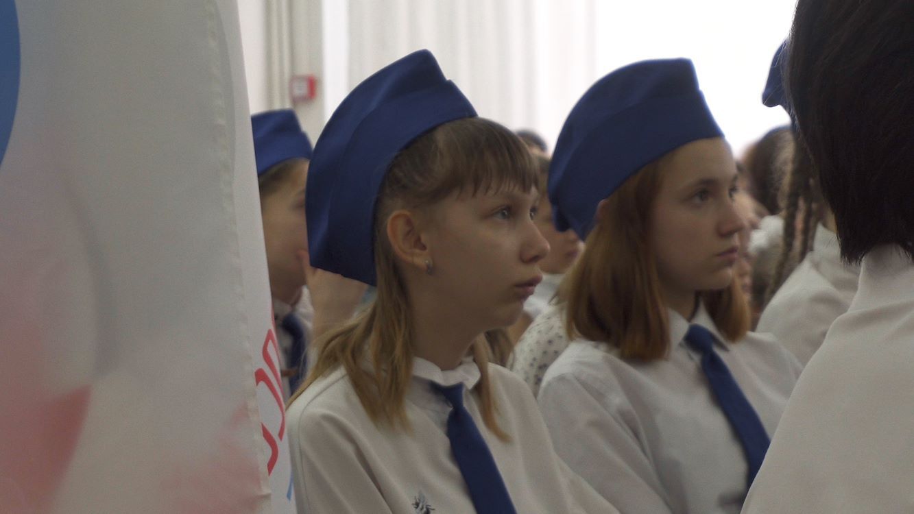 Groep leerlingen in formele kleding met blauwe hoeden, zittend tijdens een ceremonie of evenement.