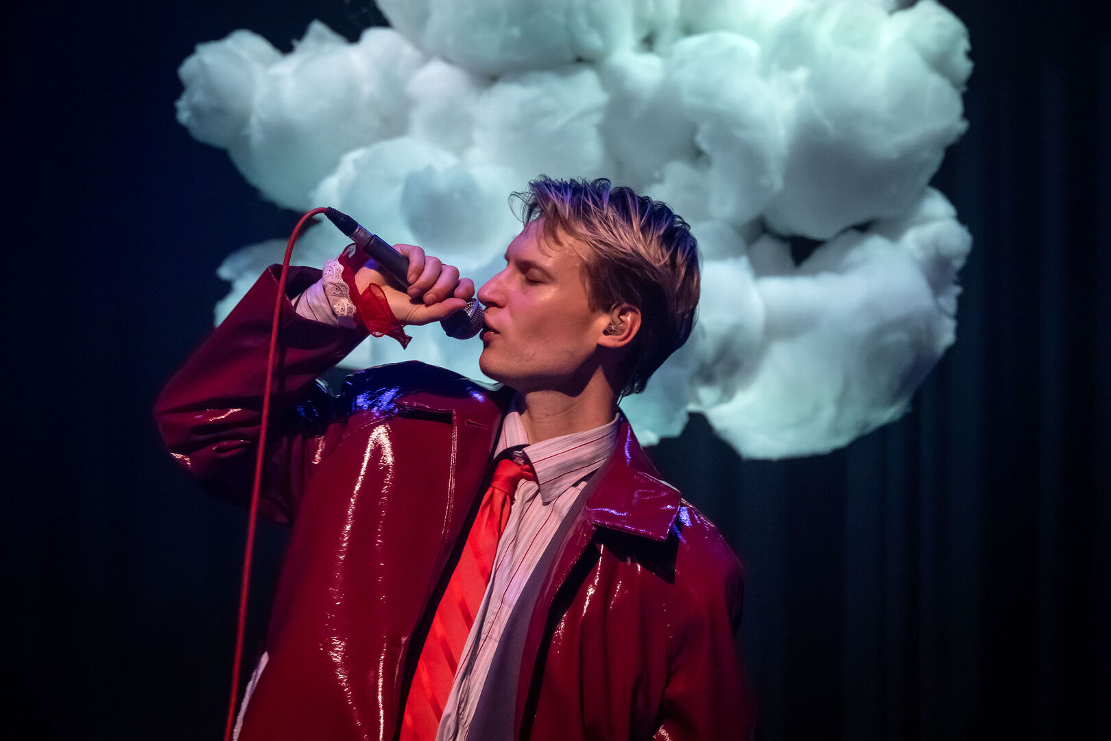 Man met een microphone in een glanzende rode jas, zingt voor een wolkenachtergrond op een podium.
