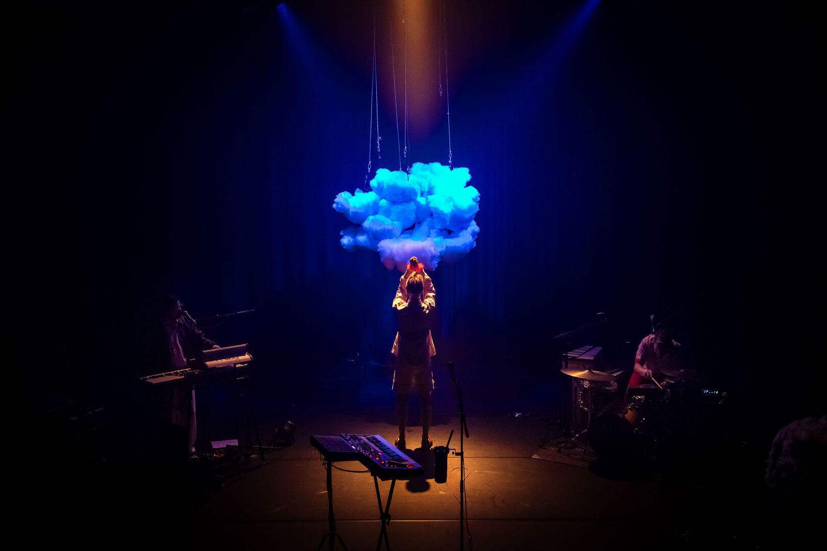 Zangeres op het podium met een wolk van licht boven zich, muzikanten aan de zijkant, sfeervolle verlichting.
