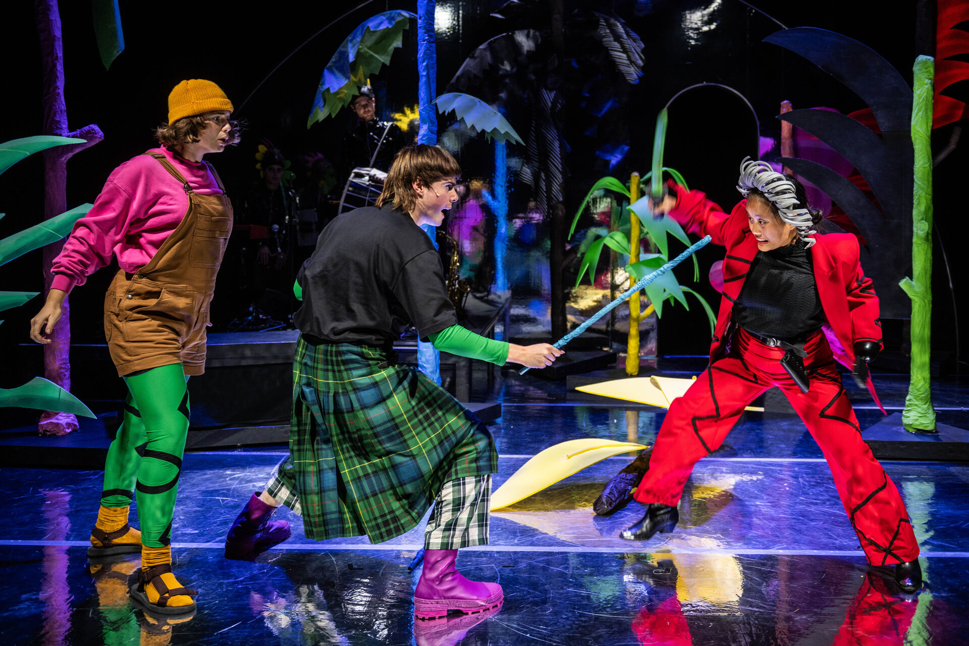 Drie performers in kleurrijke kostuums voeren een theatrale scène op een levendig decor met abstracte elementen uit.