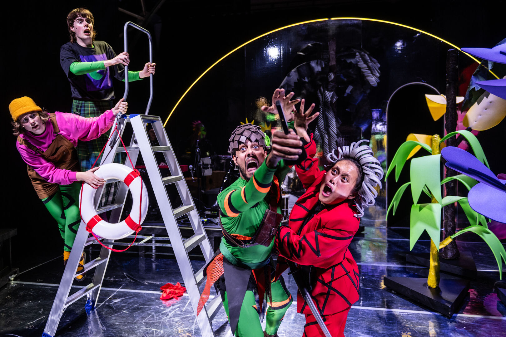 Vijf kleurrijk geklede artiesten in een circussetting voeren een act uit met een ladder en creatieve rekwisieten.