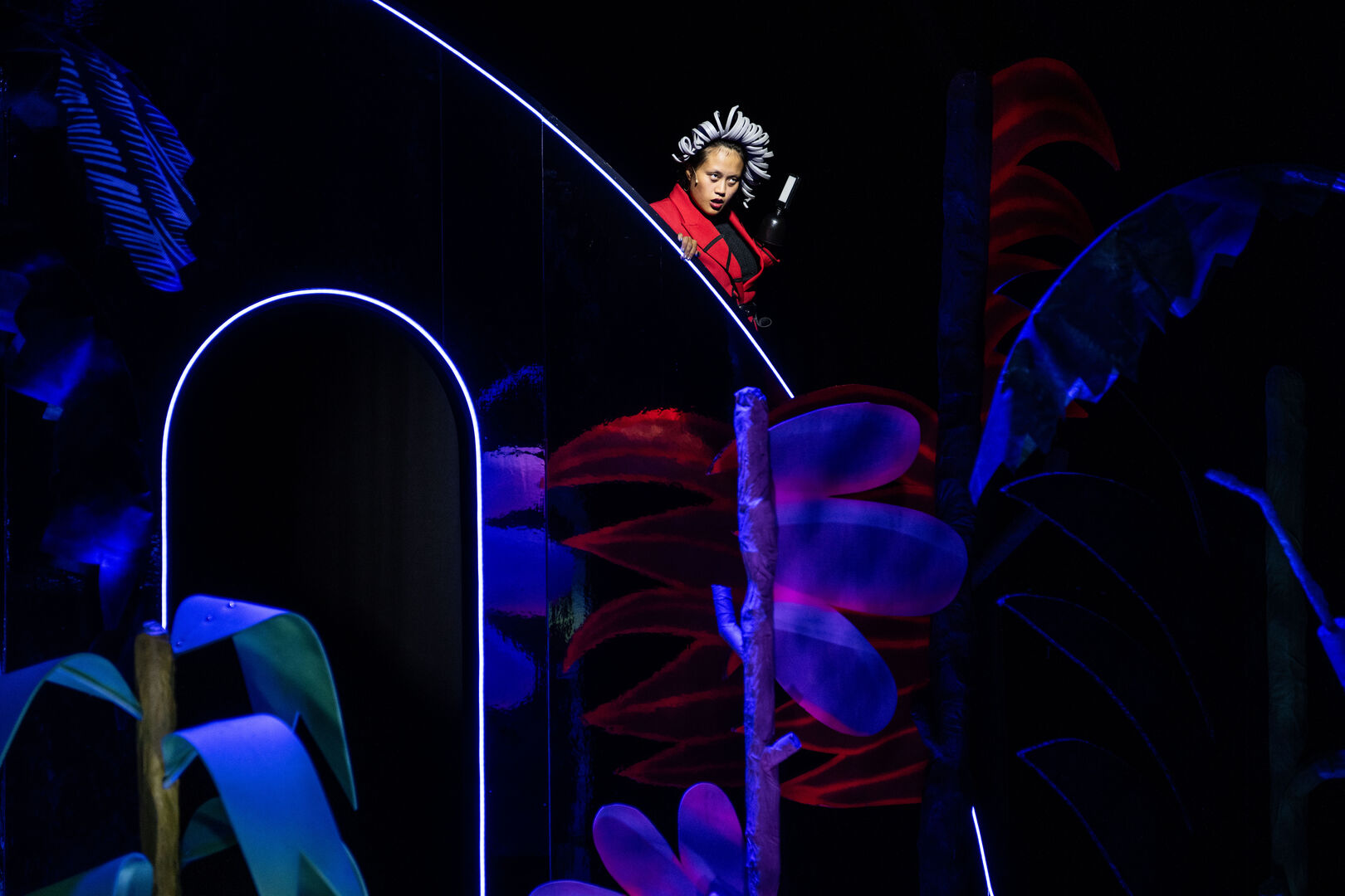 Vooraanstaande performer met kroon in een kleurrijk decor met kleurrijke planten en neonverlichting.