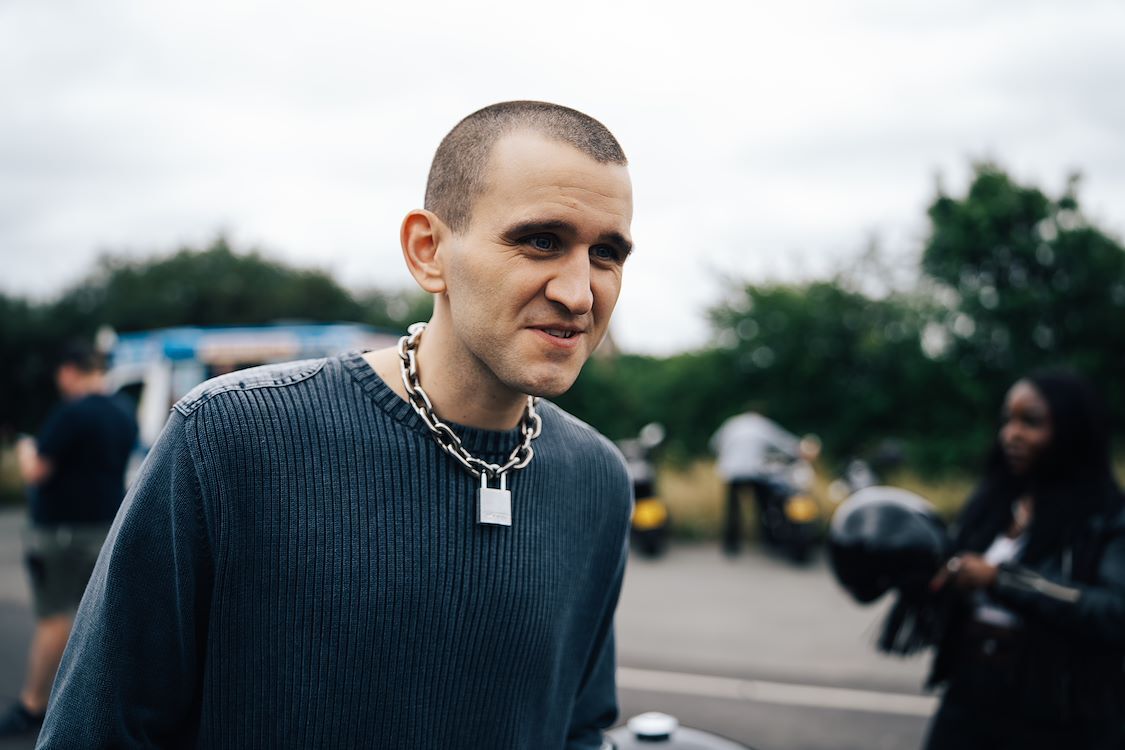 Jongeman met een kort, geschoren hoofd en een ketting, glimlacht terwijl hij buiten staat. Groene omgeving op de achtergrond.