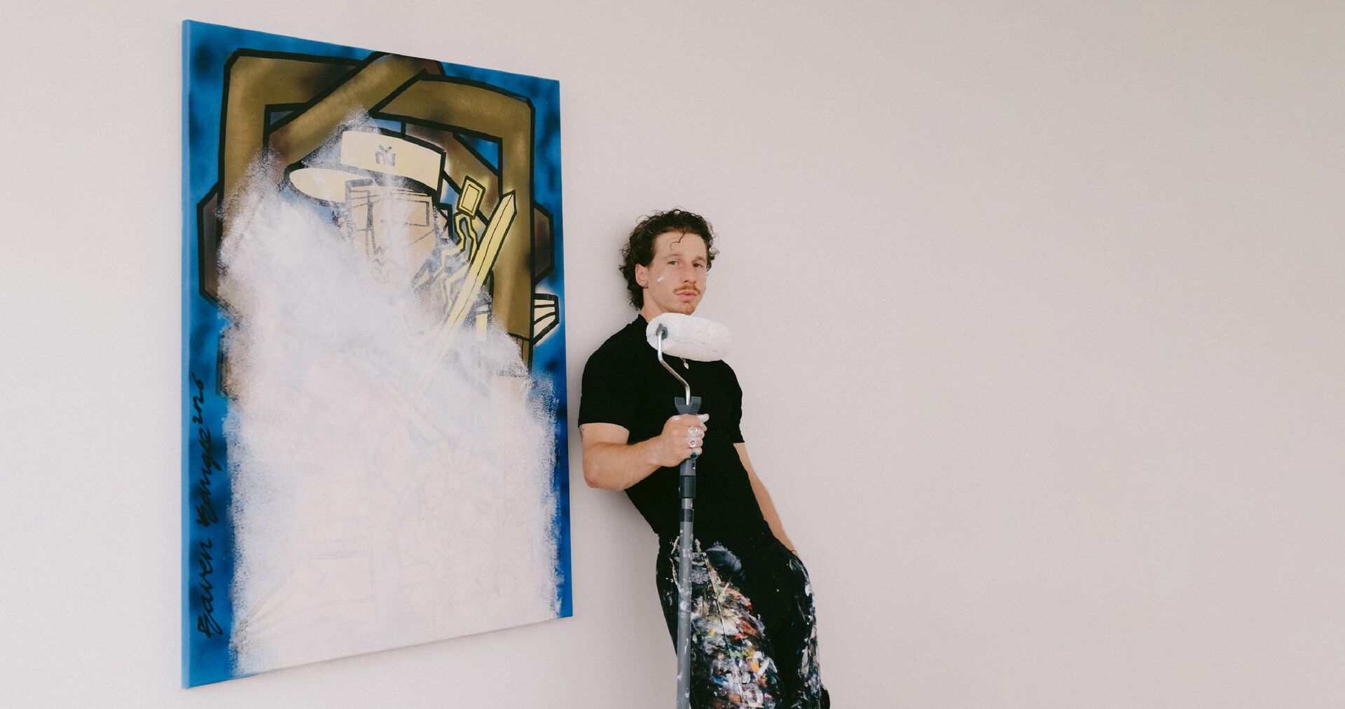 Bram Raven Kangee Ravenhorst leunt tegen een muur naast een abstracte schilderij, gekleed in een zwart shirt en met verfsporen op de broek.