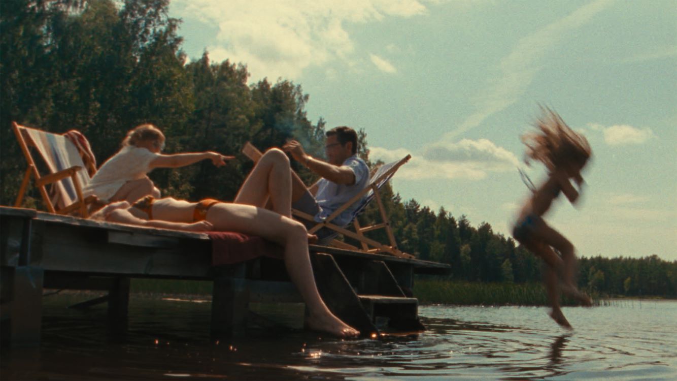 Drie kinderen spelen bij een meer; twee zitten op een steiger en één springt in het water. Zon en bomen op de achtergrond.