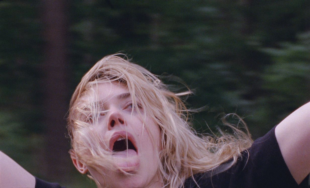 Een meisje met wapperend haar, die enthousiast en met open mond in de camera kijkt, omringd door bomen.