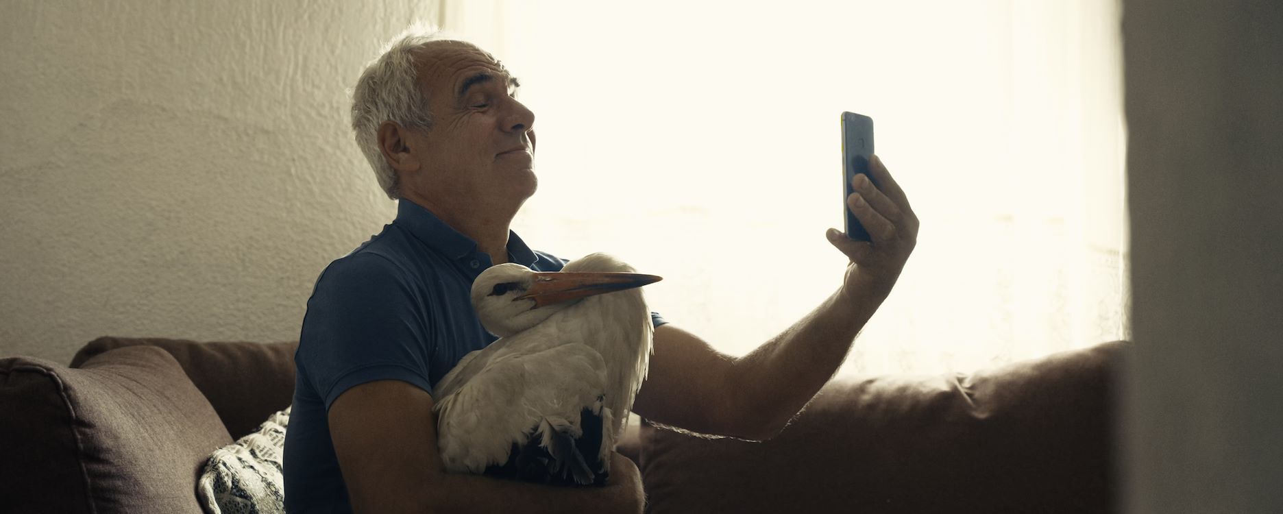 Man met een kalmeerende uitdrukking, zittend op een bank en een vogel vasthoudend, terwijl hij een selfie maakt.
