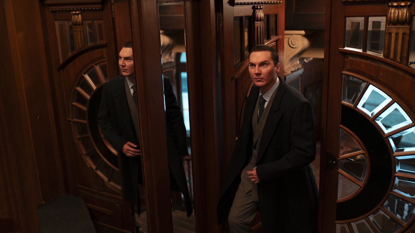 Man in een kamer met draaideur, gekleed in een net pak en kijkend naar de camera, met vintage decor op de achtergrond.