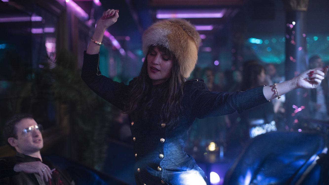 Vrouw in een feesterie draagt een opvallende hoed en danst, omringd door kleurrijke lichten en mensen.