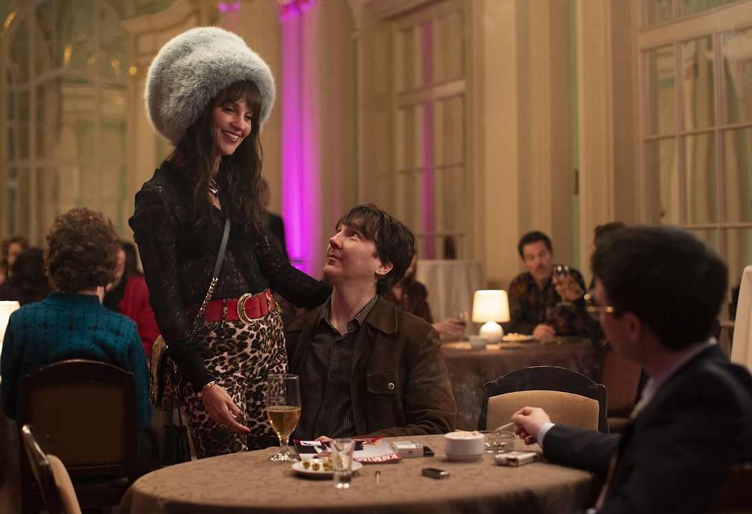 Een man en vrouw in formele kleding staan aan een tafel in een feestelijke ruimte met zachte verlichting en andere gasten.