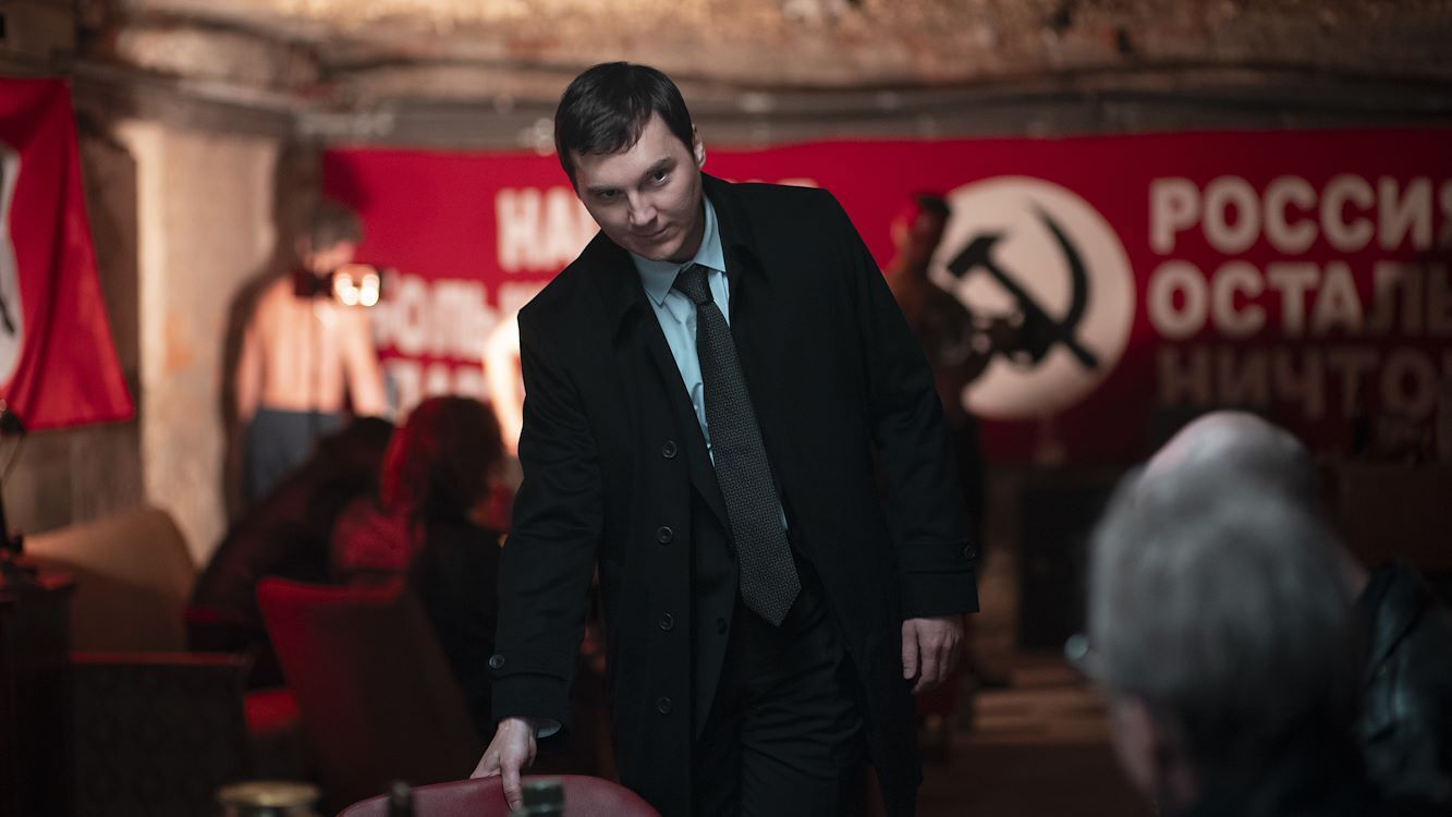 Een man in een zwart jasje staat in een ruimte met rode communistische symbolen op de achtergrond.